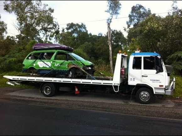 A large white and blue flat deck tow truck hauling a badly damaged green mini van