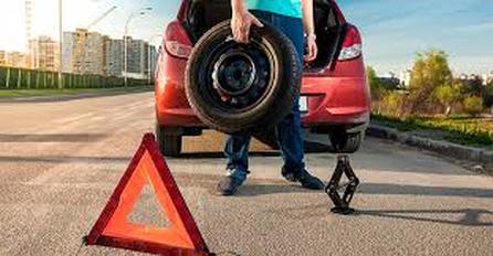 A hazard triangle on the road with a man changing a tire on a red hatchback vehicle