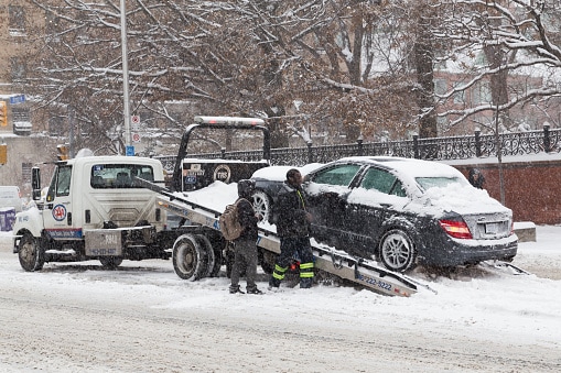 A flat deck tow truck removing a car from a city street in the winter time